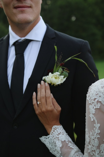 Brudens hand mot brudgummens blomsterboutonnière, ringen synlig