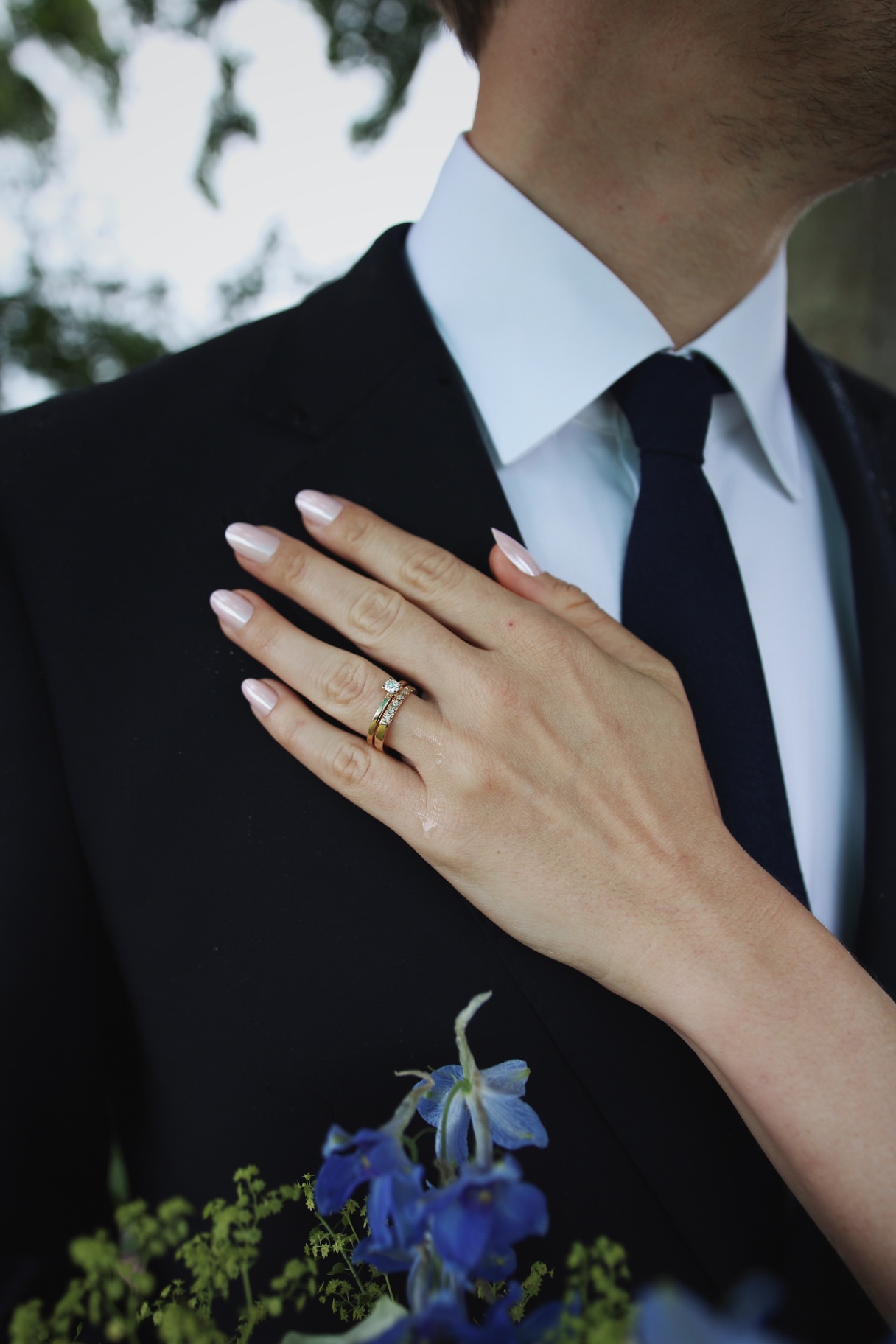 Brudens hand med vigselring vilar mot brudgummens kostym med blå delphinium